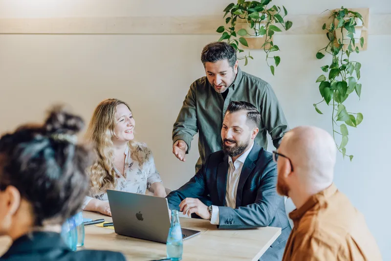 Group of colleagues having a discussion around a table with a laptop and water bottles, surrounded by hanging plants.