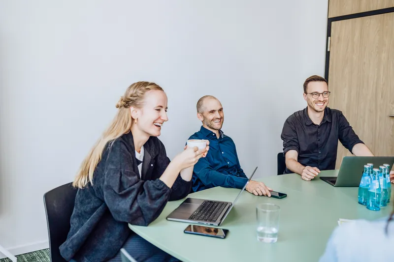 Three people sitting at a table with laptops, smiling and having a discussion in a bright office setting.