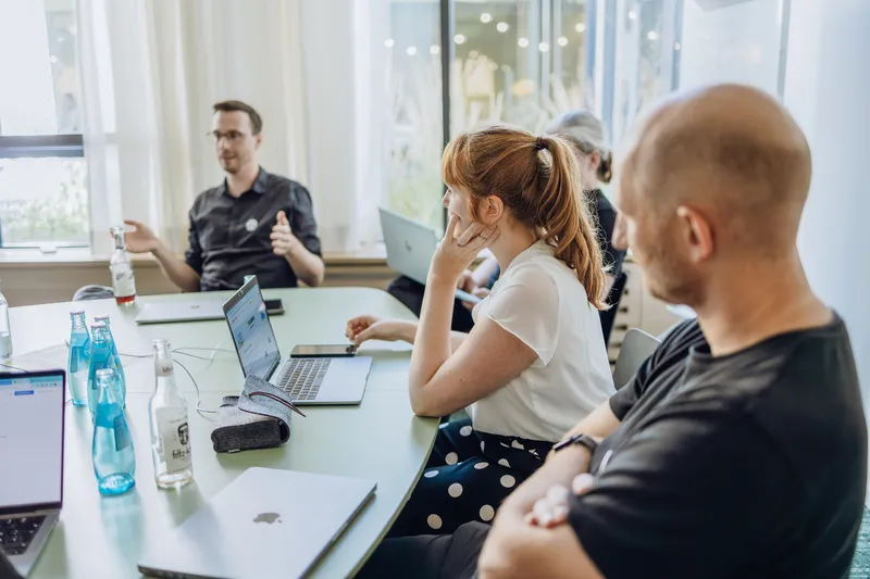 A group of people sitting around a table in a meeting room, with laptops and bottles of water on the table. One person is speaking while others listen attentively.